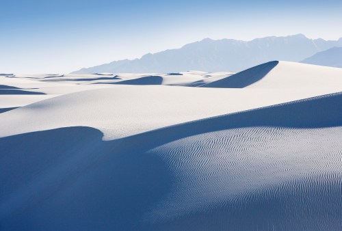 A serene landscape of white sand dunes under a clear blue sky, with distant mountains in the background.