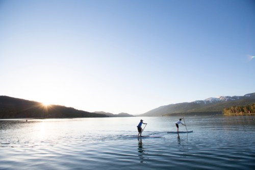 Two people paddleboarding on a calm lake at sunrise, surrounded by mountains and clear blue skies.