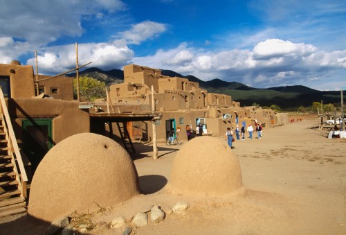 A traditional adobe village with rounded structures, people walking, and mountains in the background under a cloudy sky.