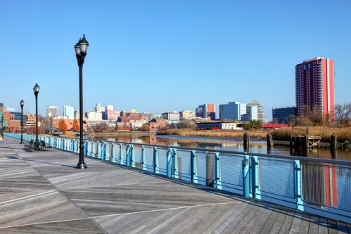 A waterfront view of a city skyline with buildings, a river, and a wooden boardwalk under a clear blue sky.