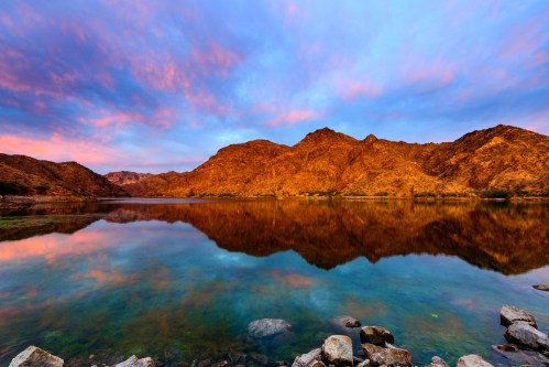 A serene lake reflects colorful clouds and rocky mountains at sunset, creating a tranquil landscape scene.