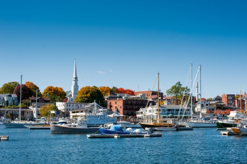 A scenic harbor with boats, colorful autumn trees, and a church steeple against a clear blue sky.