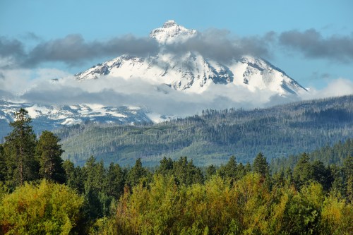 Snow-capped mountain peak surrounded by clouds, with lush green forests in the foreground.
