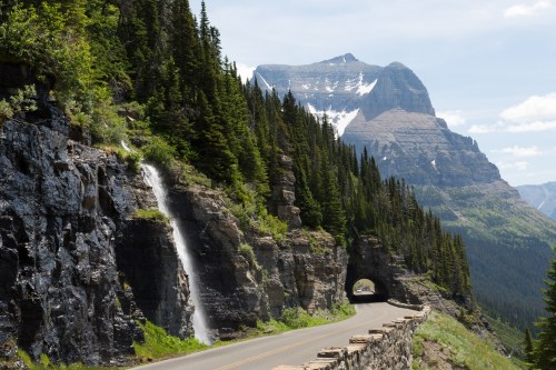 A scenic mountain road with a waterfall and a tunnel, surrounded by lush green trees and towering peaks.