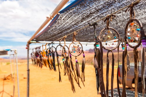 Colorful dreamcatchers hanging under a shaded canopy, set against a desert landscape with blue skies.