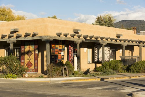 Adobe-style building with colorful quilts hanging on the walls, surrounded by greenery and a clear blue sky.