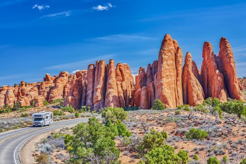 An RV drives along a road beside towering red rock formations under a clear blue sky.