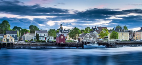 A serene waterfront scene featuring colorful houses, a docked boat, and a cloudy sky reflecting on the water.