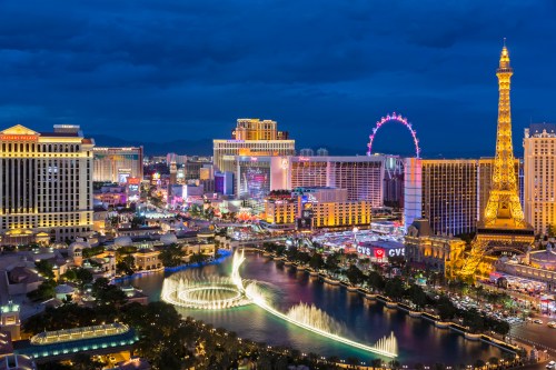 A vibrant night view of the Las Vegas Strip, featuring the Eiffel Tower replica and colorful lights reflecting on water.