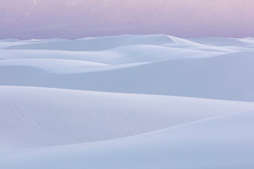 A serene landscape of smooth, undulating white sand dunes under a soft pastel sky.