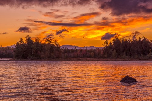 A serene lake at sunset, with vibrant orange and purple skies reflecting on the water and silhouetted trees in the foreground.