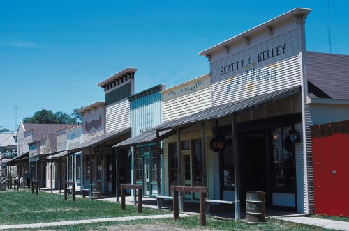 A row of historic storefronts with colorful facades under a clear blue sky. Green grass in the foreground.