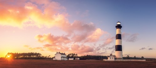 A striped lighthouse stands beside a white house at sunset, with colorful clouds and a serene landscape.