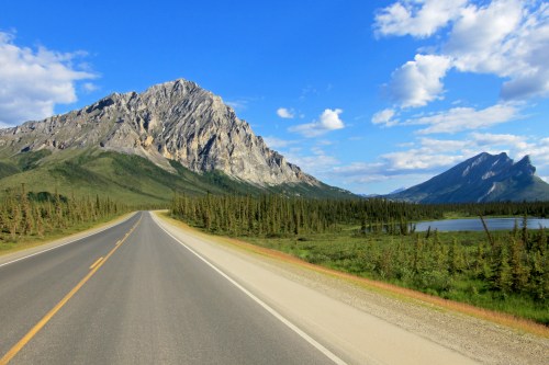 A scenic road winds through lush greenery, flanked by mountains and a lake under a blue sky with scattered clouds.