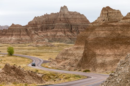A winding road through rugged, colorful rock formations and grassy plains under a cloudy sky.