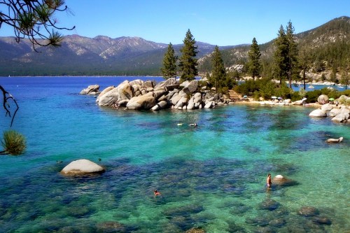 A serene lake with clear turquoise water, rocky shores, and pine trees, set against a backdrop of mountains.