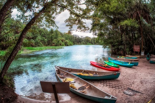 Canoes lined up by a serene river surrounded by lush greenery and trees under a cloudy sky.