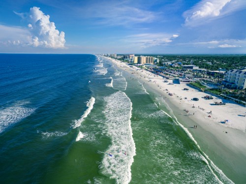 Aerial view of a beach with waves, people, and buildings along the shore under a blue sky with clouds.