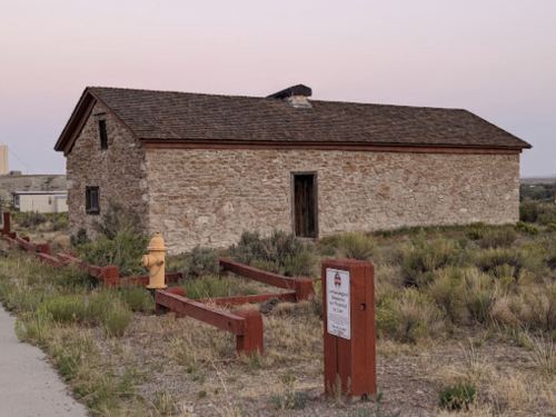 A stone building with a wooden roof, surrounded by grass and a wooden fence, at dusk. A fire hydrant is nearby.