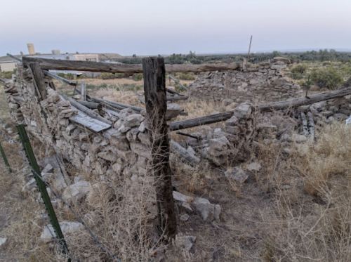 Ruins of a stone structure surrounded by dry grass and wooden posts, set against a distant landscape at dusk.