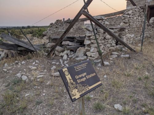 A weathered sign for Granger Stage Station stands near the ruins of a stone building at sunset.