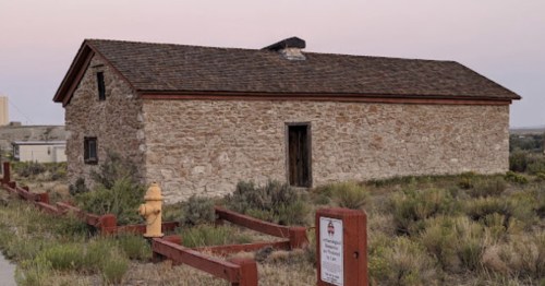 A rustic stone building with a sloped roof, surrounded by grass and shrubs, near a fire hydrant and a sign.