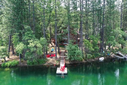 Aerial view of a serene lakeside cabin surrounded by tall trees, with a dock and red chairs by the water.