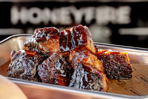 A close-up of glazed, smoked meat chunks on a tray, with a blurred background featuring the word "HONEYFIRE."