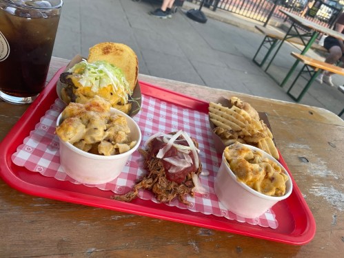 A red tray with pulled pork, mac and cheese, and waffle fries, alongside a drink, on a wooden table outdoors.