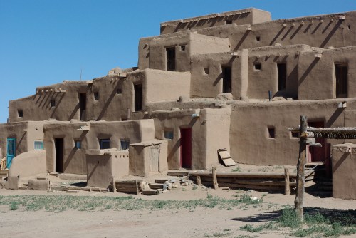 Adobe-style buildings with flat roofs and colorful doors, set against a clear blue sky. Green grass in the foreground.