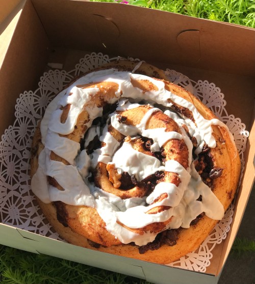 A large cinnamon roll with cream cheese frosting in a decorative box, resting on a lace doily.