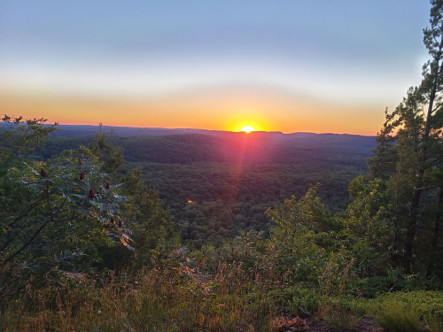 A vibrant sunset over a lush green landscape, with trees framing the view and warm colors in the sky.