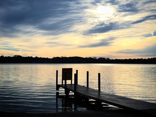 A serene lake at sunset, with a wooden dock extending into the water and clouds reflecting in the calm surface.