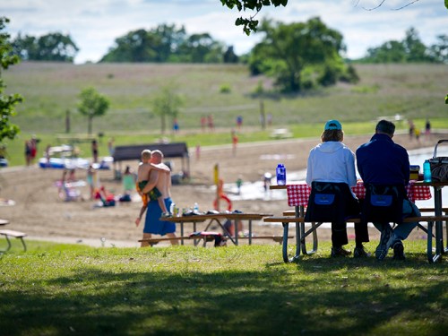 Two people sit at a picnic table, while others enjoy a beach area in the background on a sunny day.