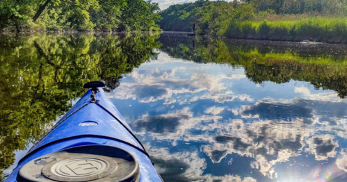 A blue kayak on calm water reflects trees and clouds in a serene natural setting.