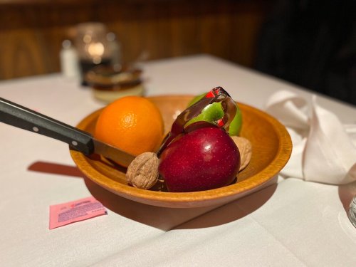 A wooden bowl with an orange, green apple, red apple, and walnuts, with a knife resting on the edge.