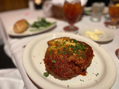 A plate of breaded chicken topped with marinara sauce and herbs, served with a roll and a drink in the background.