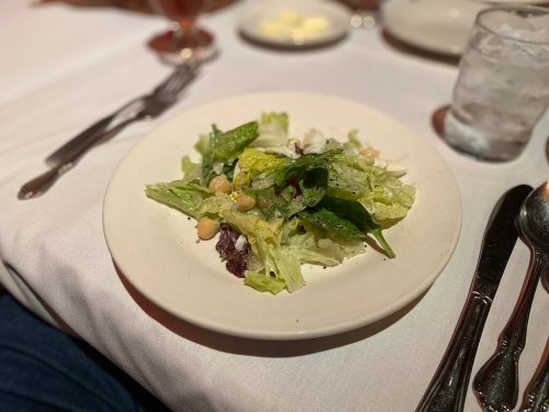 A fresh salad with greens, chickpeas, and dressing on a white plate, set on a table with utensils and a glass of water.