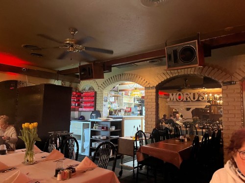 Interior of a restaurant featuring tables with white tablecloths, a bar area, and a kitchen visible in the background.