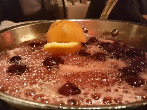 A close-up of a bubbling drink with cherries and an orange slice in a metal bowl, set in a restaurant ambiance.