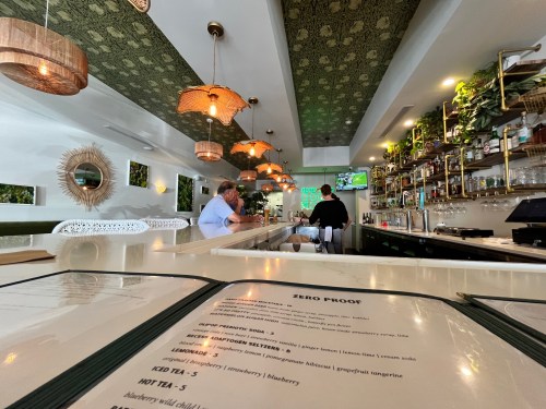 A modern bar interior with green decor, pendant lights, and a menu on the counter. Two patrons are seated at the bar.