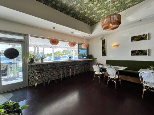 Bright restaurant interior with green accents, bar seating, and decorative plants by large windows.