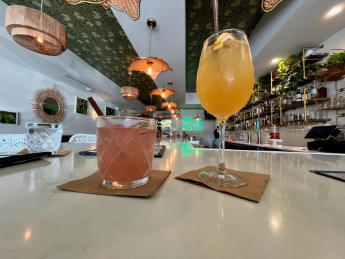 Two colorful cocktails on a bar counter, with a stylish interior featuring hanging lights and greenery in the background.