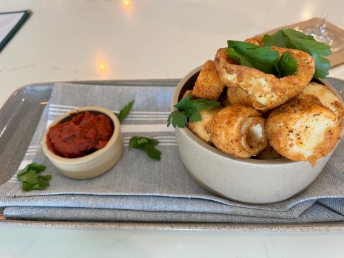 A bowl of crispy fried snacks garnished with parsley, served with a side of red sauce on a gray napkin.