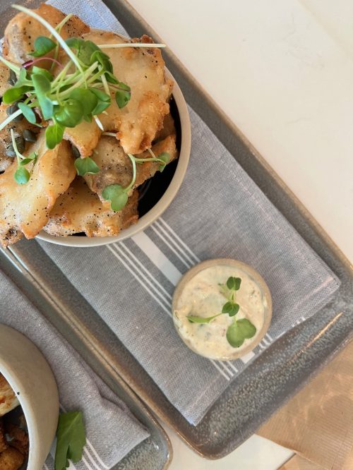 A bowl of crispy fried food topped with microgreens, alongside a small dish of creamy sauce on a gray tray.
