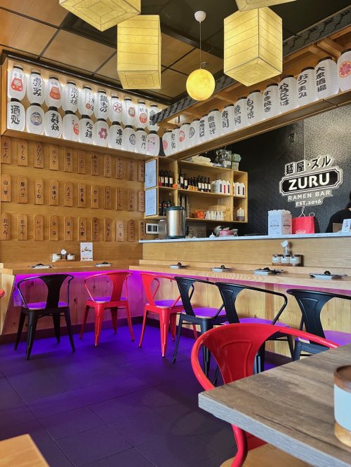 Interior of a ramen bar with wooden walls, colorful chairs, hanging lanterns, and Japanese decor.