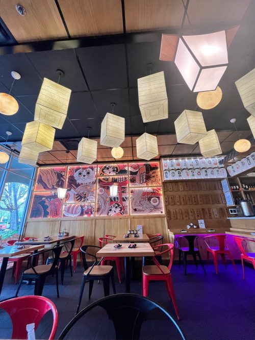 Interior of a modern restaurant featuring wooden decor, colorful lanterns, and red chairs around tables.