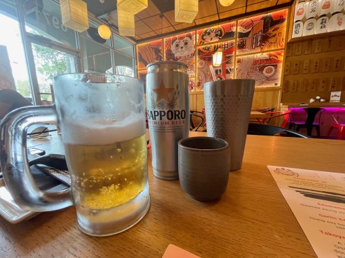 A glass of beer, a can of Sapporo, and two cups on a wooden table in a cozy restaurant setting.