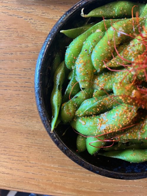 A bowl of edamame sprinkled with seasoning, placed on a wooden table.