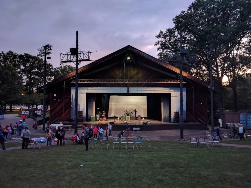 An outdoor stage with a band setup, surrounded by people and empty chairs, under a cloudy sky at dusk.